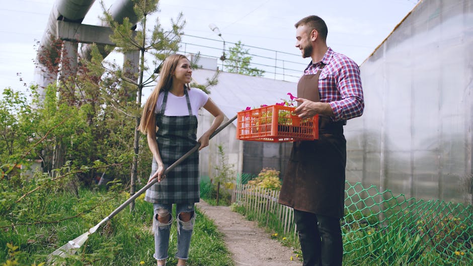 A young woman with long hair, wearing a white t-shirt and a plaid apron over ripped jeans, stands in a lush garden holding a rake. She is smiling and engaging in garden maintenance activities. Opposit
