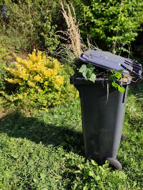A black wheeled garden waste bin positioned on a grassy area during daylight, filled with a mix of garden clippings including dried grasses, green leaves, and plant stems. The bin is located in a lush
