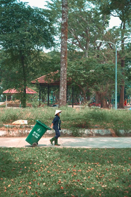 A black wheeled garden waste bin positioned on a grassy area during daylight, filled with a mix of garden clippings including dried grasses, green leaves, and plant stems. The bin is located in a lush